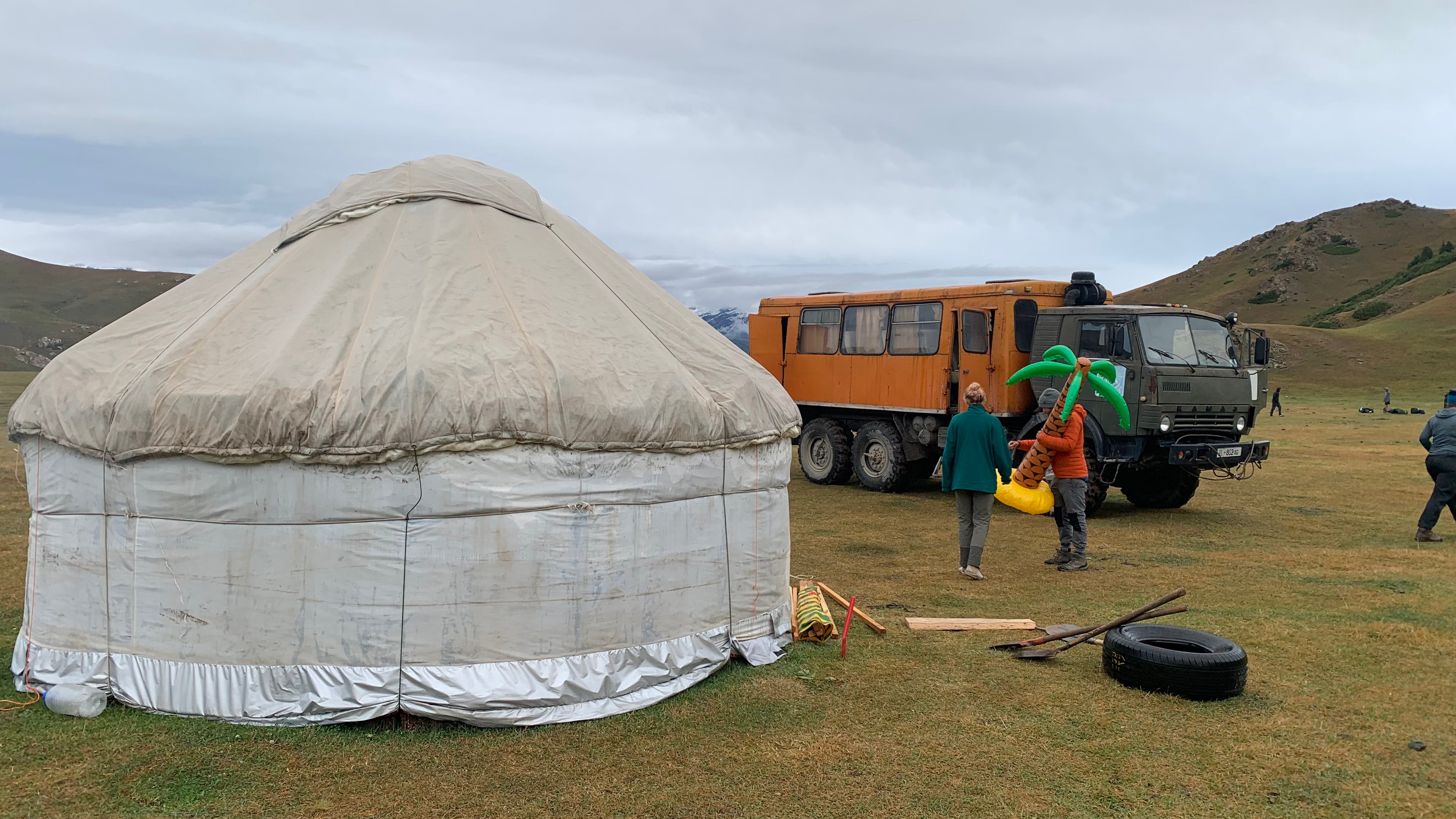 Traditional felt yurt and an orange expedition truck on open grassland beneath rolling hills