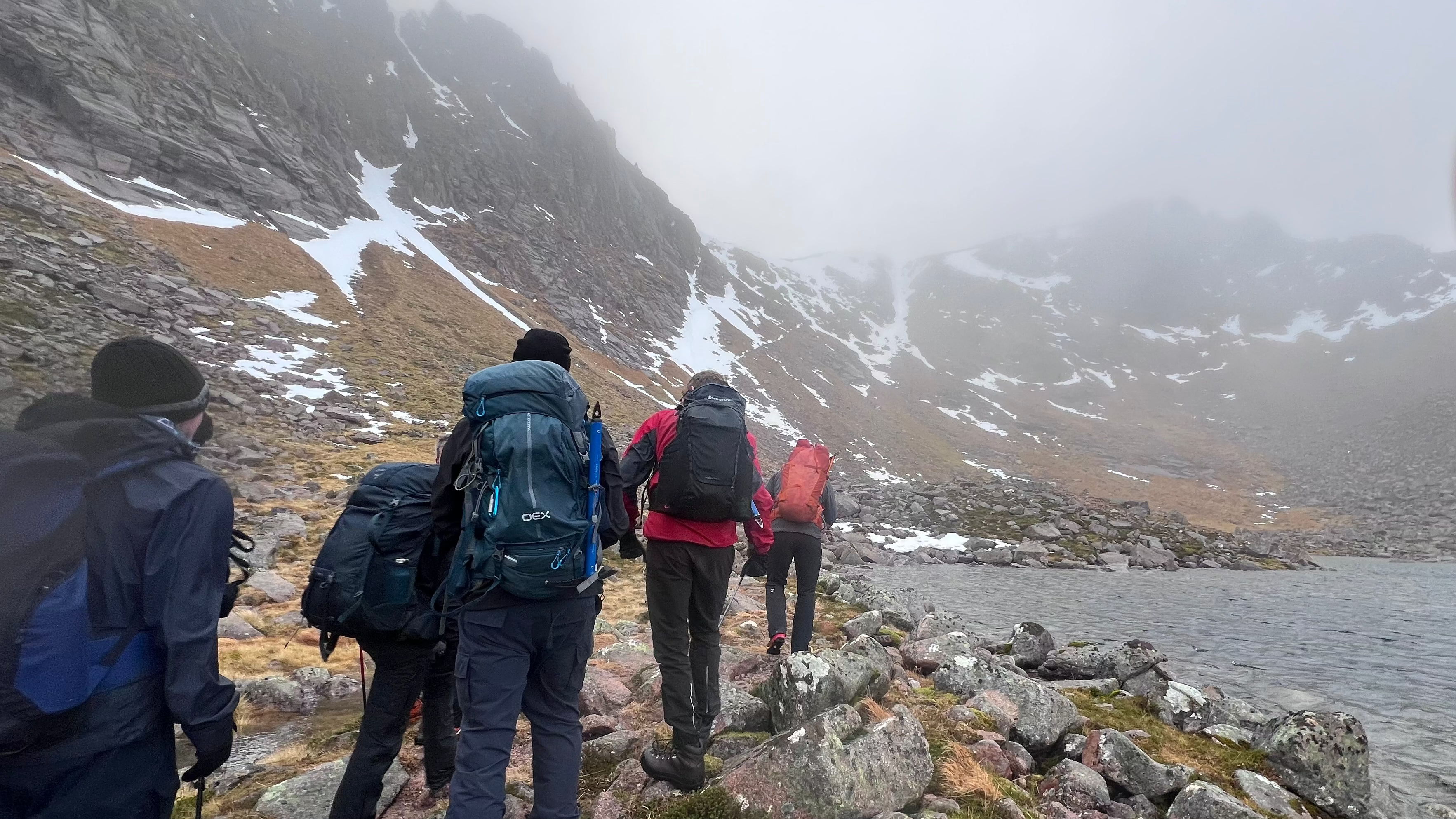 Group of hillwalkers with rucksacks skirting a misty mountain loch beneath snow-streaked crags